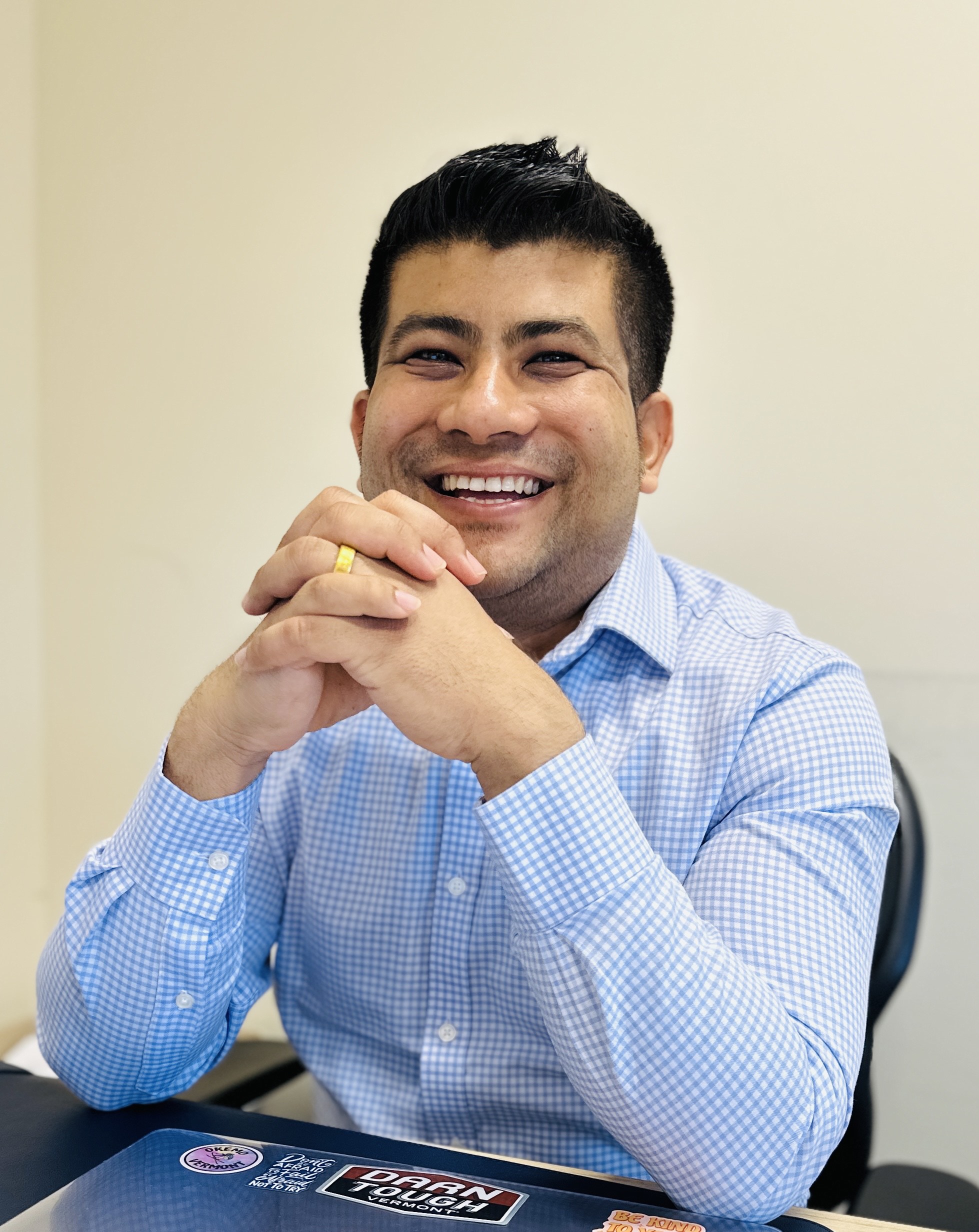 male, dark hair, blue shirt, at a desk, hands folded in front of him.  Big smile