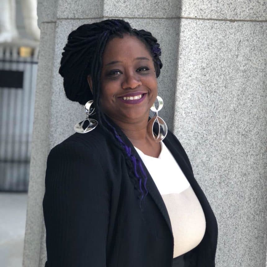 woman of color with black braids, large silver earrings, big smile, black coat with white top under it.