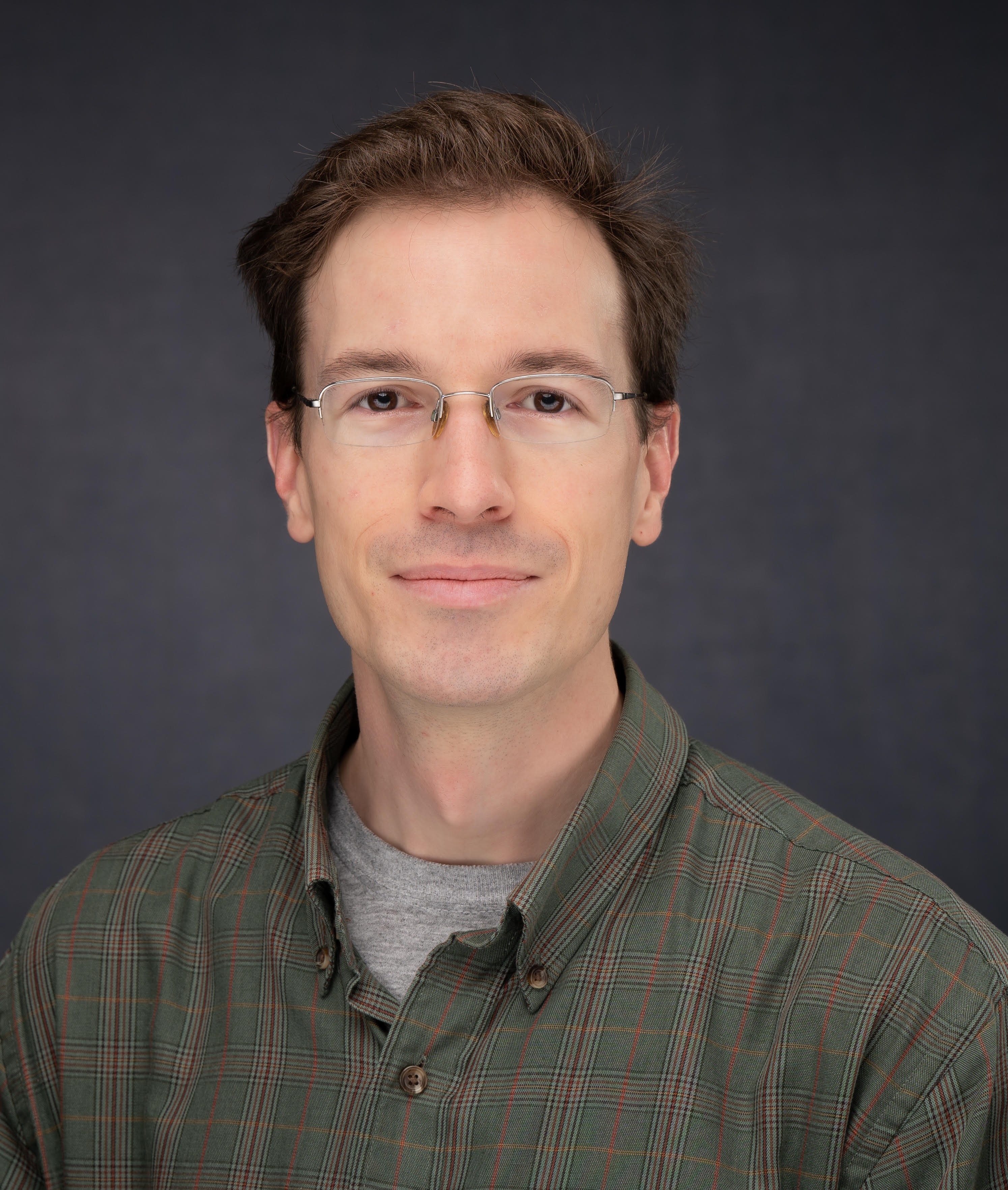 young white man, dark hair, dark plaid shirt, light glasses