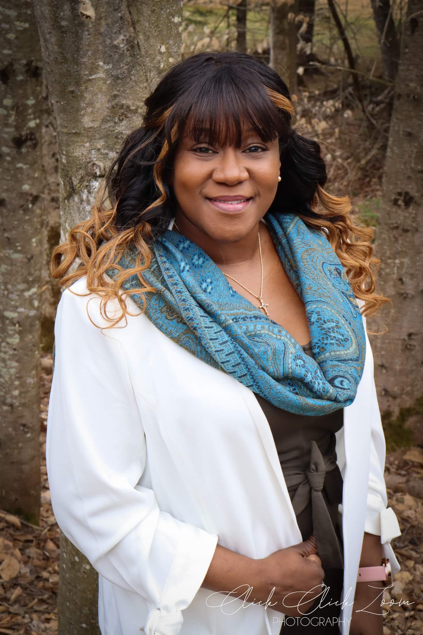 woman of color standing against a tree.  long white coat, brown shirt and blue paisley scarf