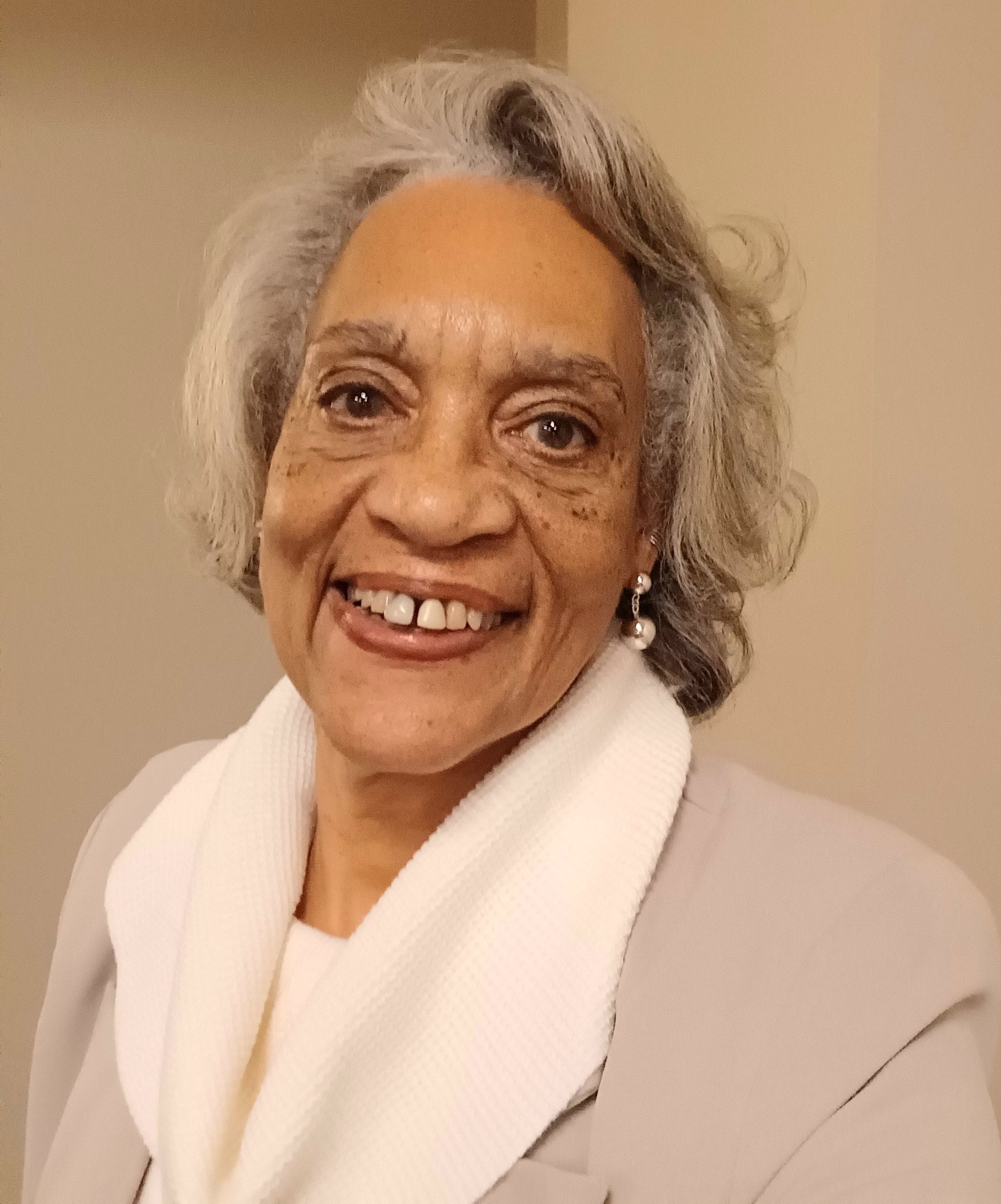 woman of color with short silver hair, brown eyes, white scarf over tan coat