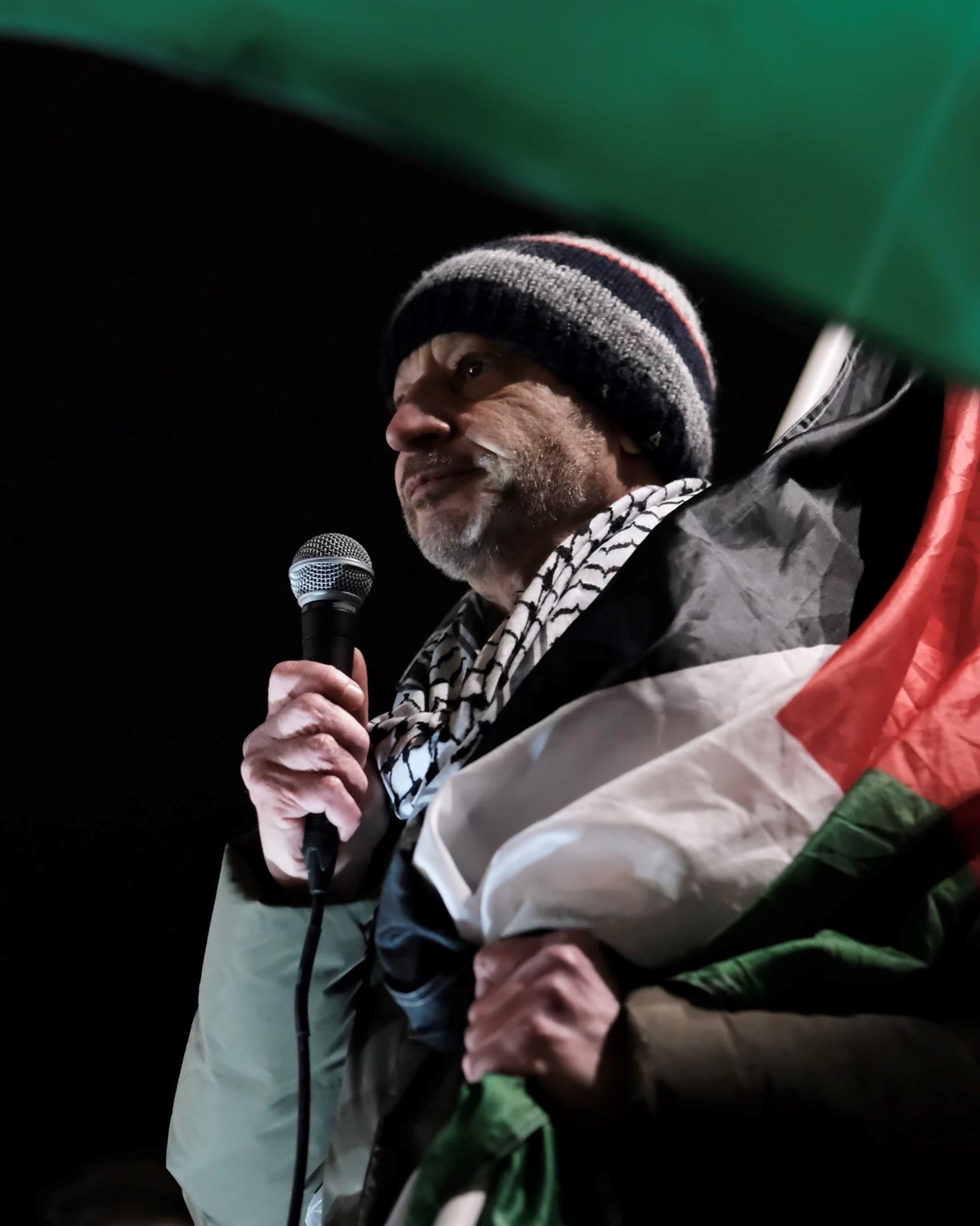 Man with microphone, wool had, black and white scarf, flag draped around back